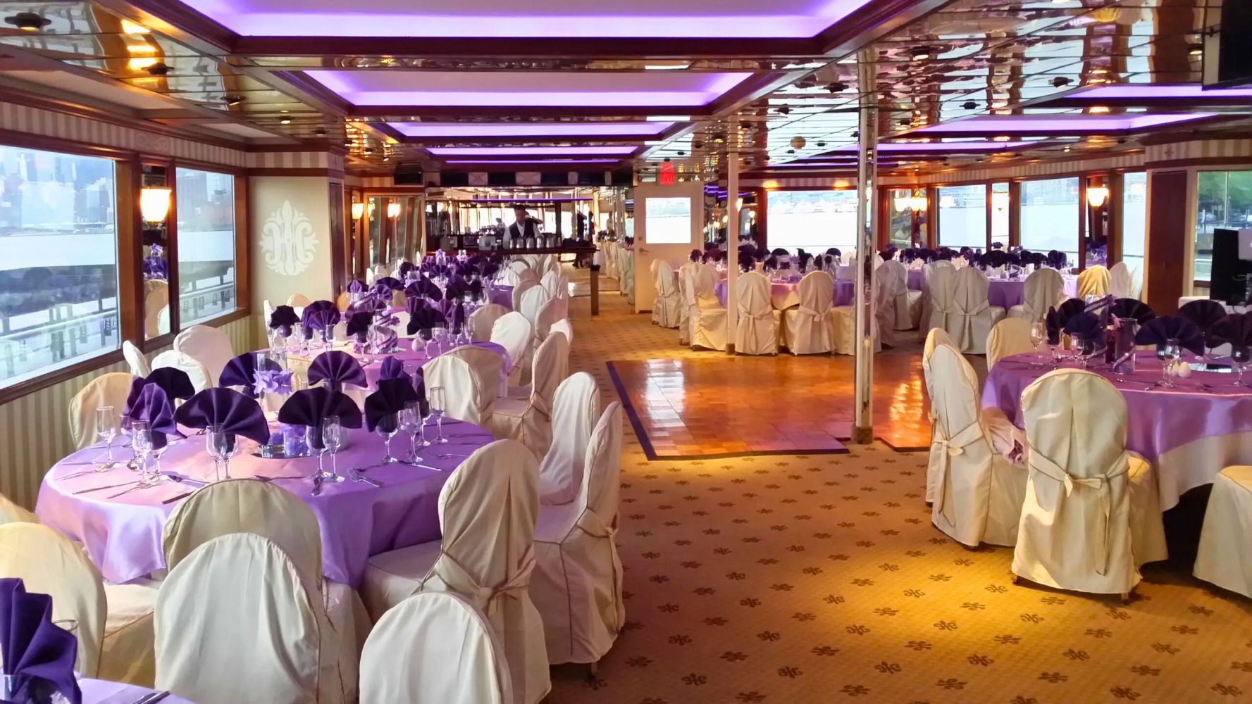 Elegant banquet hall with purple-lit ceiling and round tables set with purple napkins and white chair covers for a private party cruise