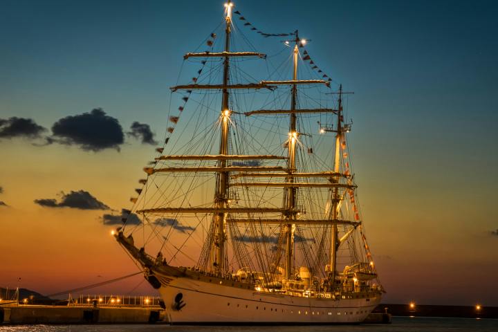 Illuminated tall ship docked at sunset with colorful sky and few clouds.