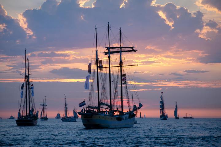 Tall ships sailing at sunset, with colorful sky and scattered clouds on the horizon.